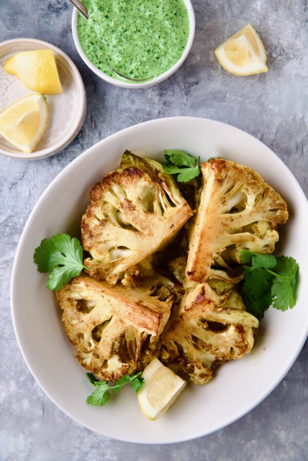 Four wedges of Curry Roasted Cauliflower Wedges with Cilantro Chutney in white bowl with lemon wedges.