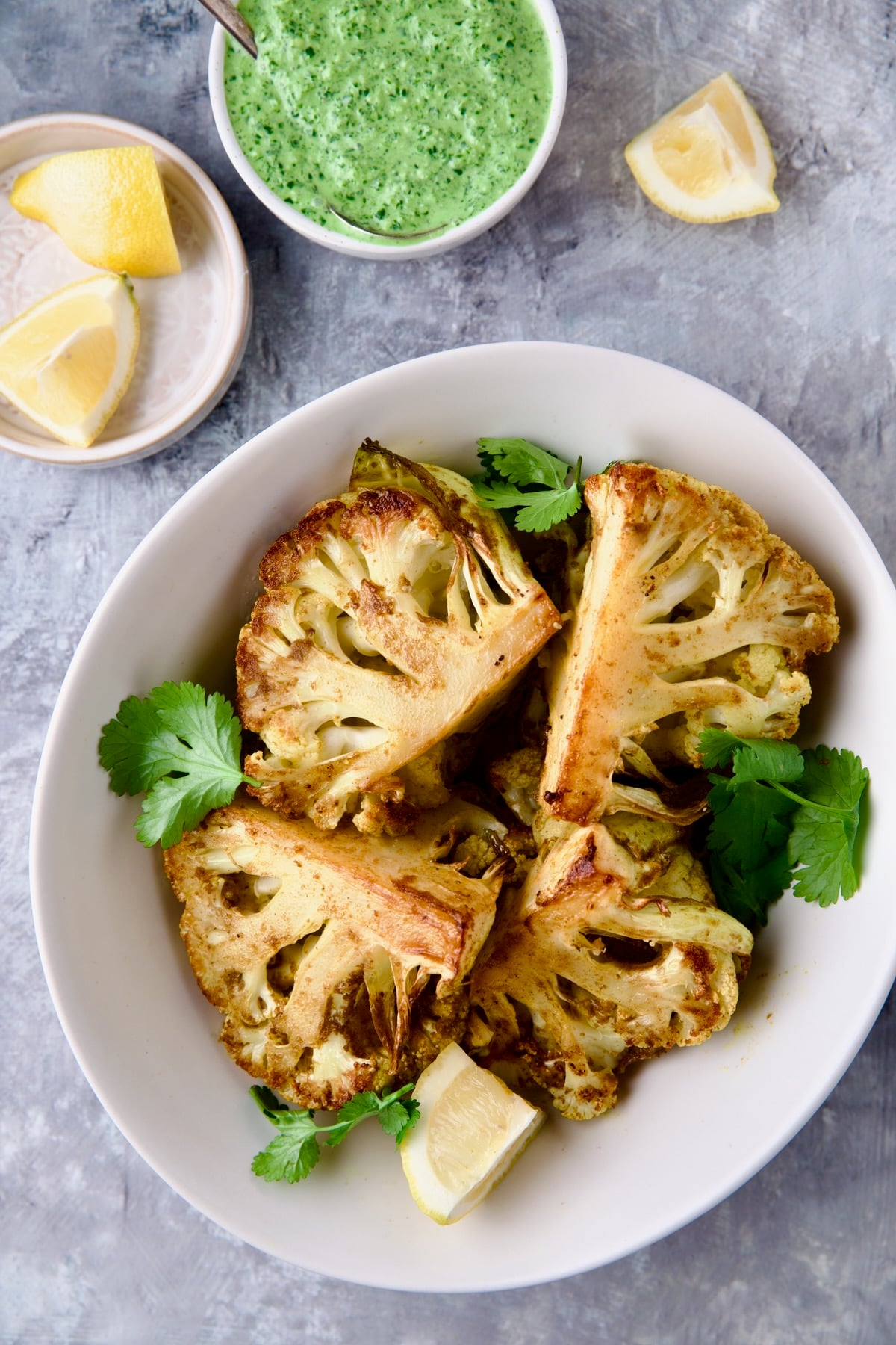 Four wedges of Curry Roasted Cauliflower Wedges with Cilantro Chutney in white bowl with lemon wedges.