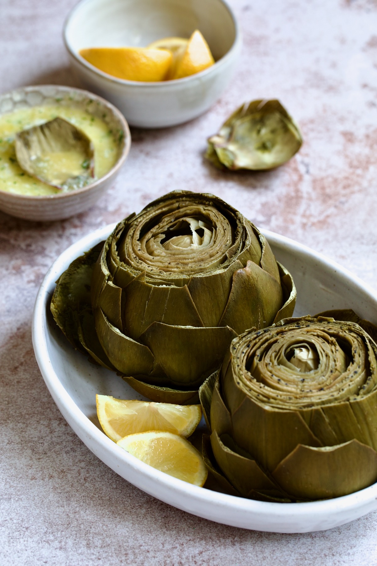 Instant Pot Artichokes in white oval serving bowl with garlic butter and lemon wedges in the background.