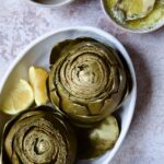 Instant Pot Artichokes (With Garlic Butter) in white oval serving bowl with lemon wedges.