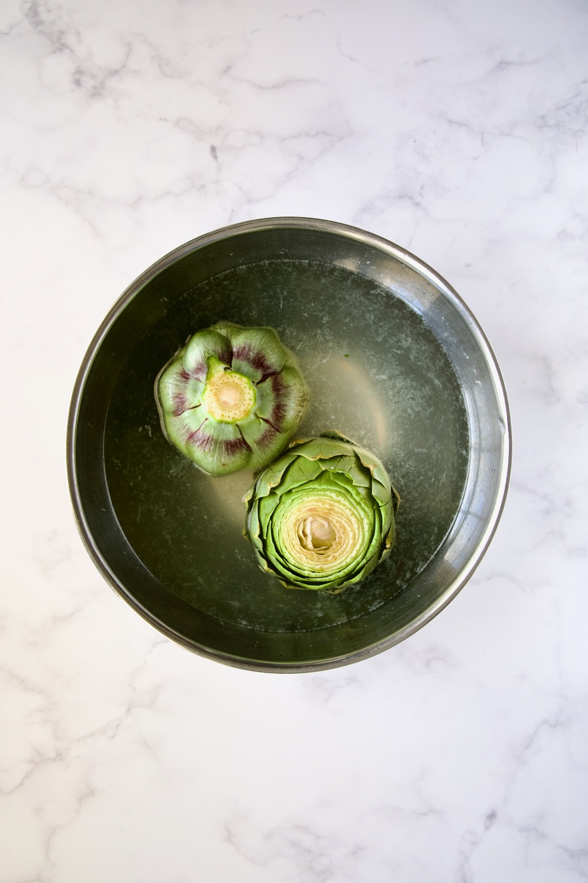 Two artichokes in lemon water in stainless steel bowl.