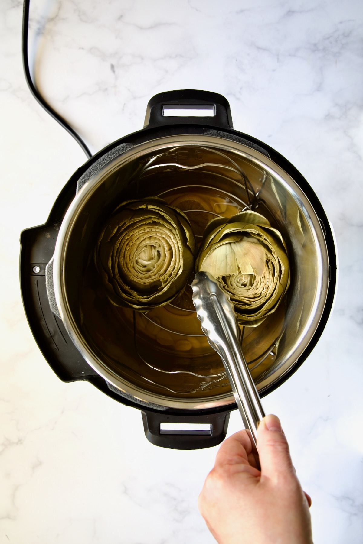 Two cooked artichokes in Instant Pot with tongs pulling one of the leaves out.