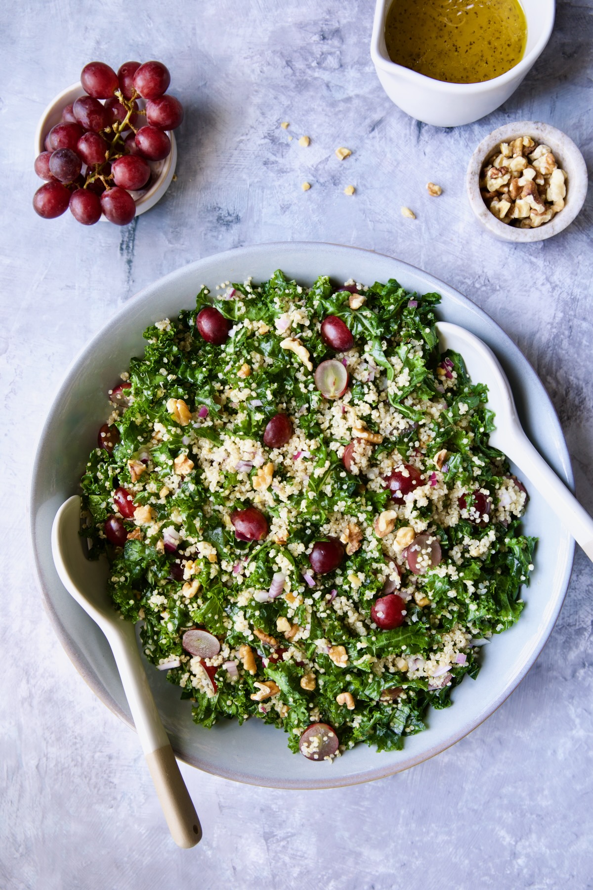Kale Quinoa Salad with Lemon Honey Dressing in gray serving bowl with serving utensils and grapes, dressing and walnuts in separate bowls.