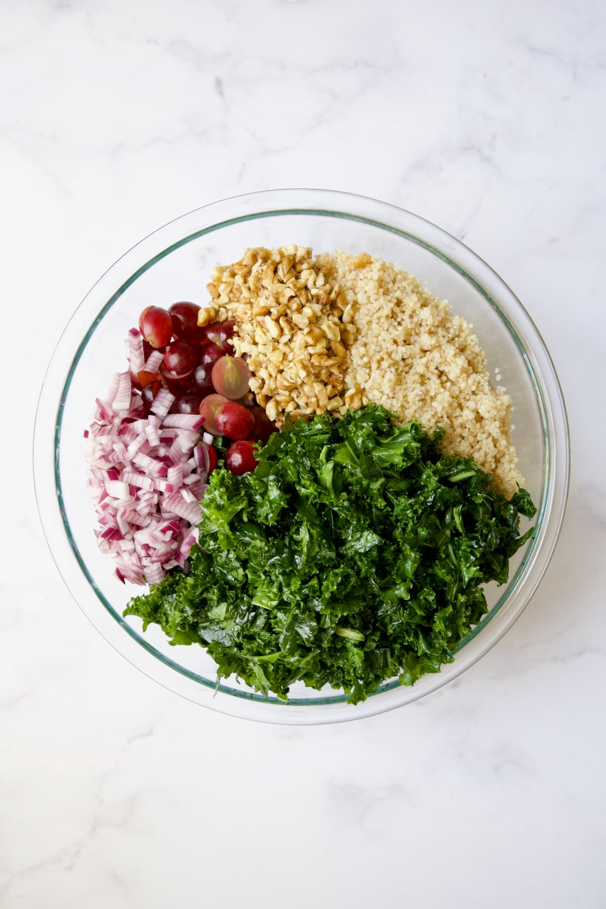 Kale, quinoa, walnuts, red grapes and red onion in glass bowl before being mixed together.
