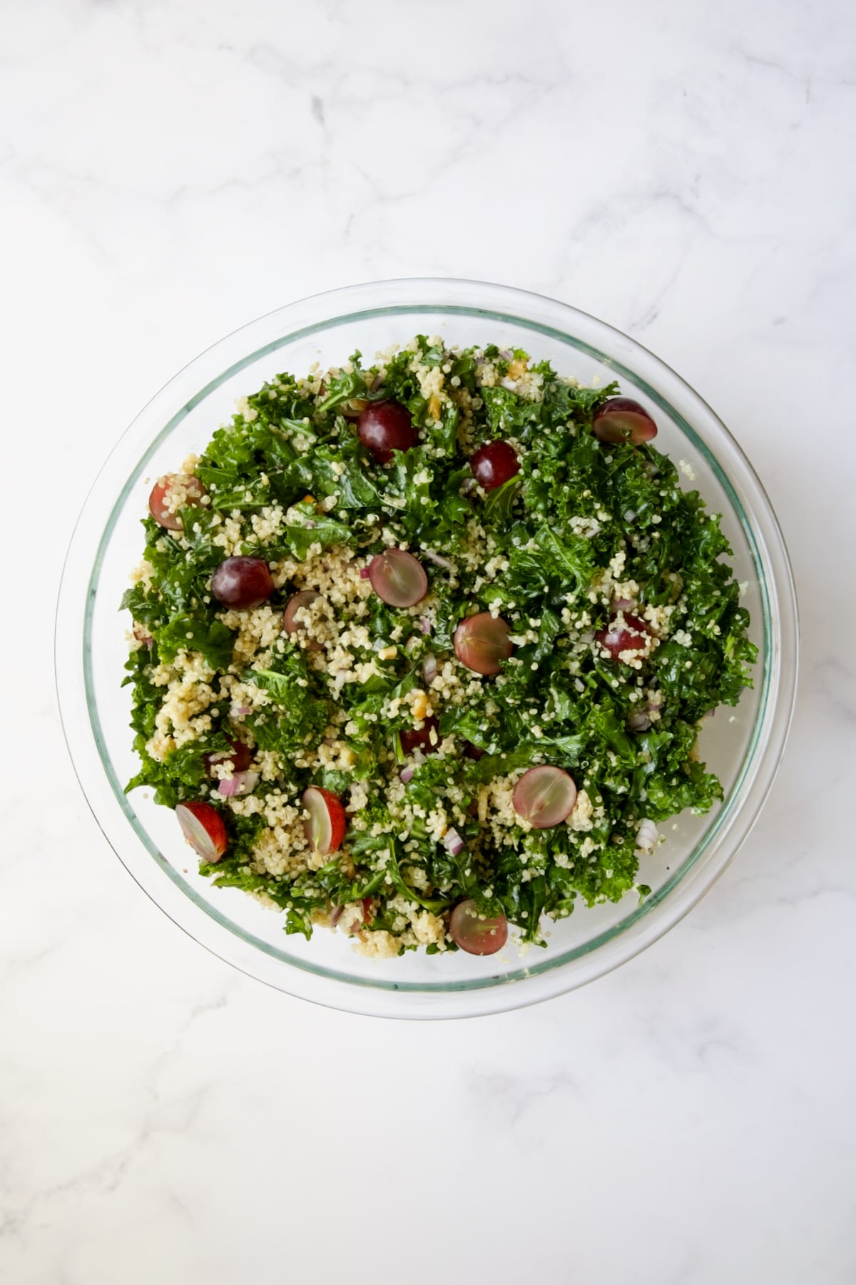 Kale, quinoa, walnuts, red grapes and red onion in glass bowl all mixed together.