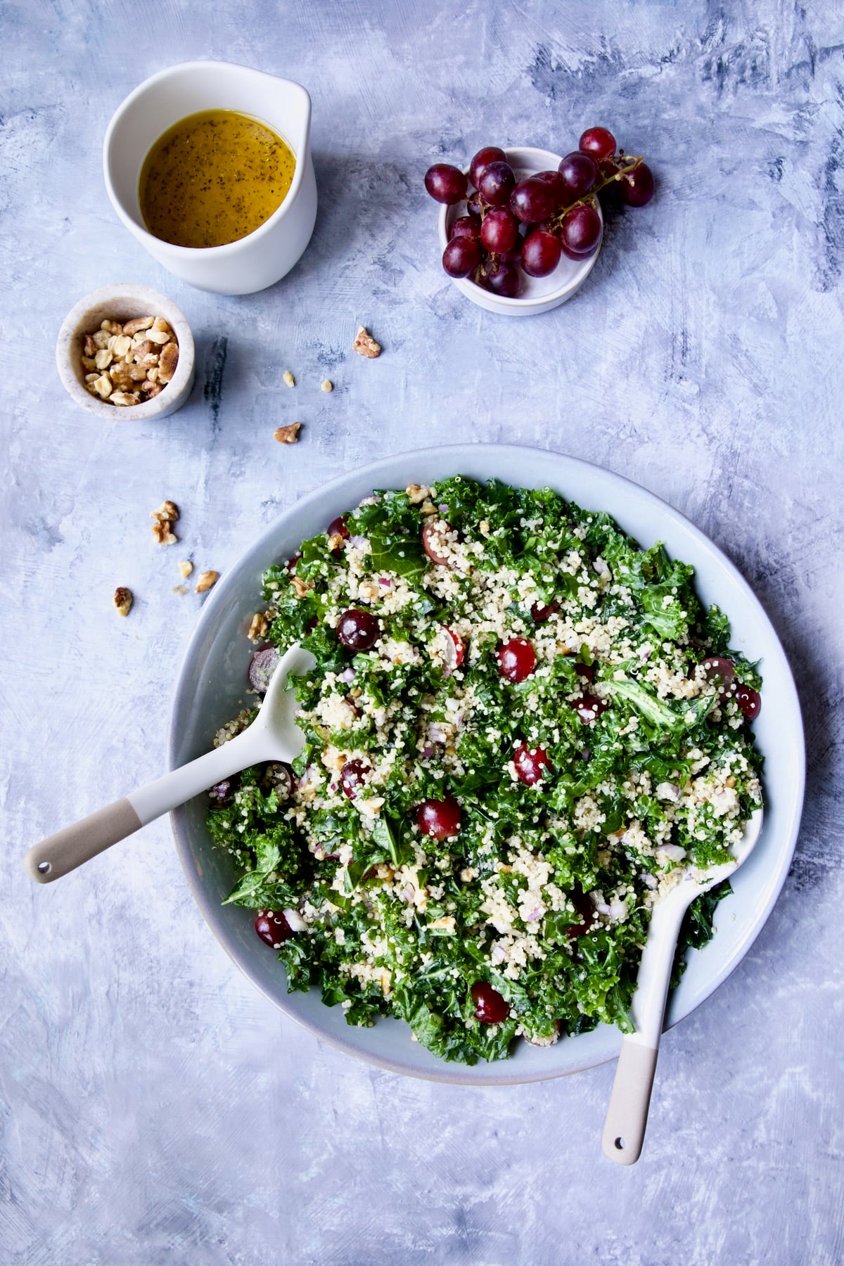 Kale Quinoa Salad with Lemon Honey Dressing in serving bowl on blue background with walnuts, dressing and grapes in separate bowls.