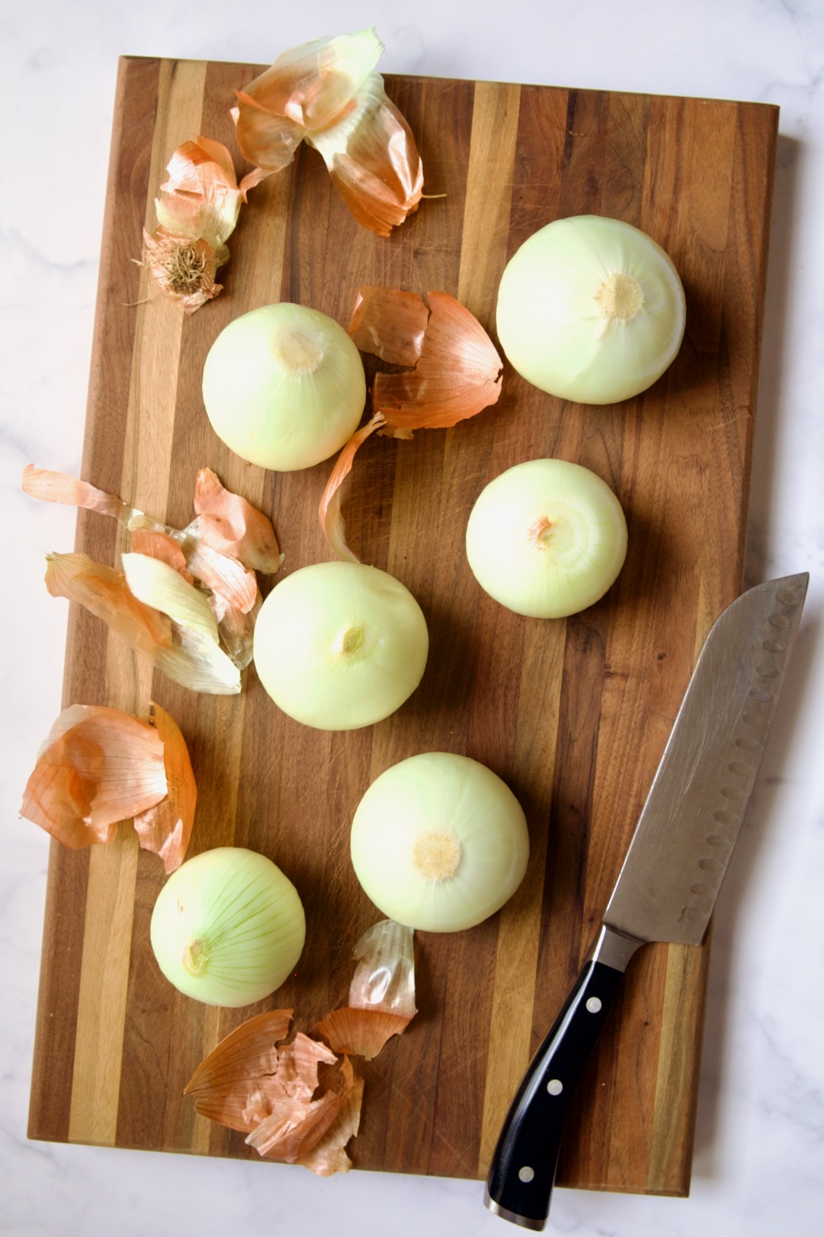 Six peeled onions with some of the peels and chef knife on wood cutting board.