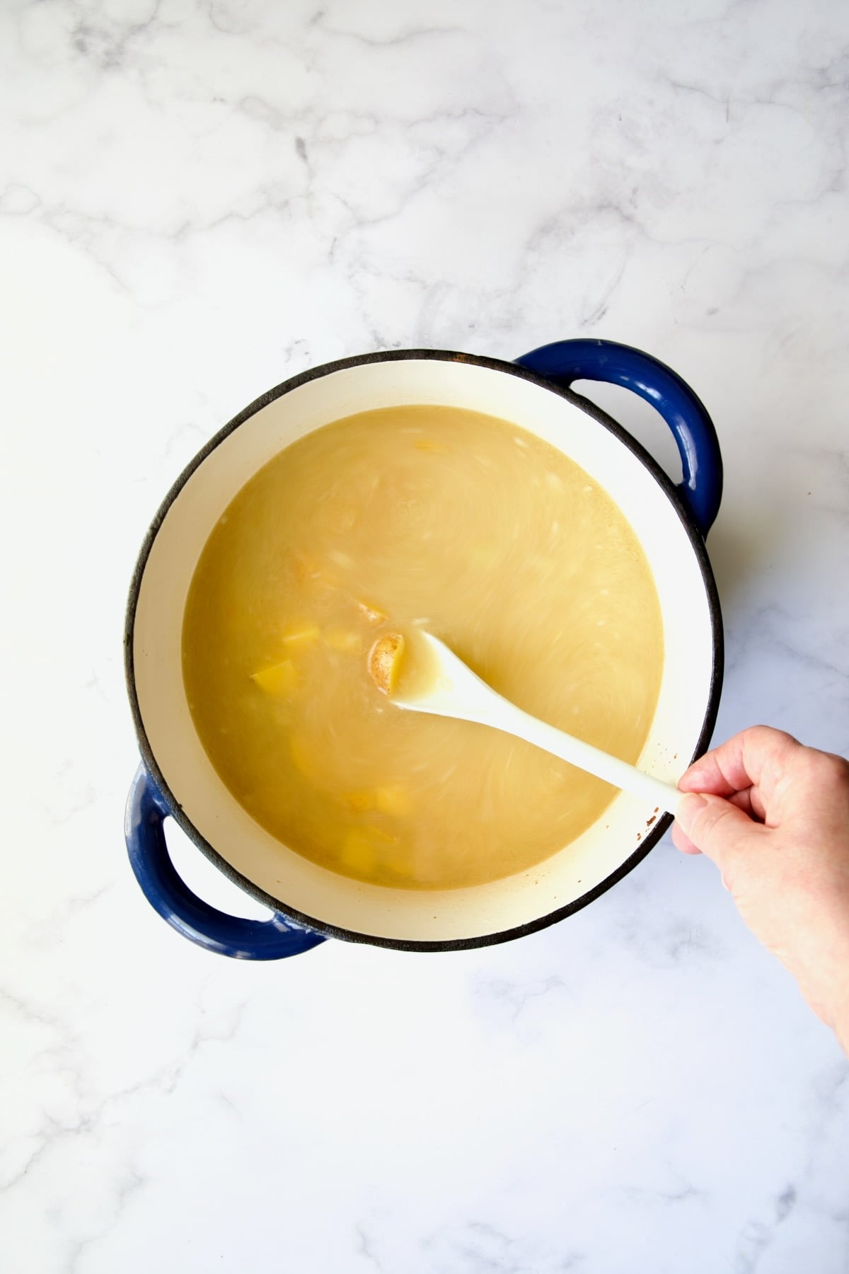 Chicken broth and potatoes added to other ingredients in blue Dutch oven with hand stirring the soup with white mixing spoon.