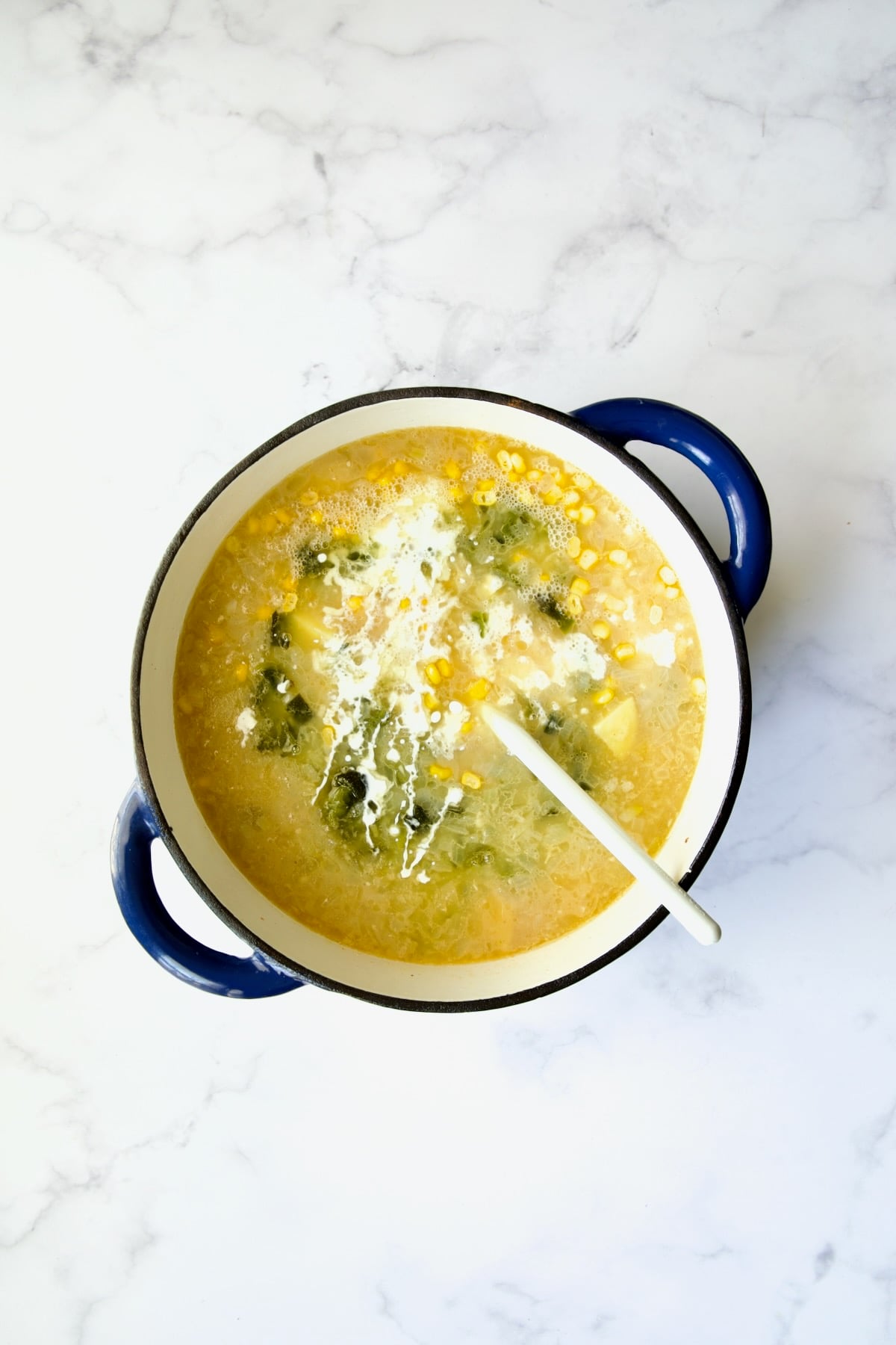 Corn, roasted poblanos and cream added to blue Dutch oven with white mixing spoon.