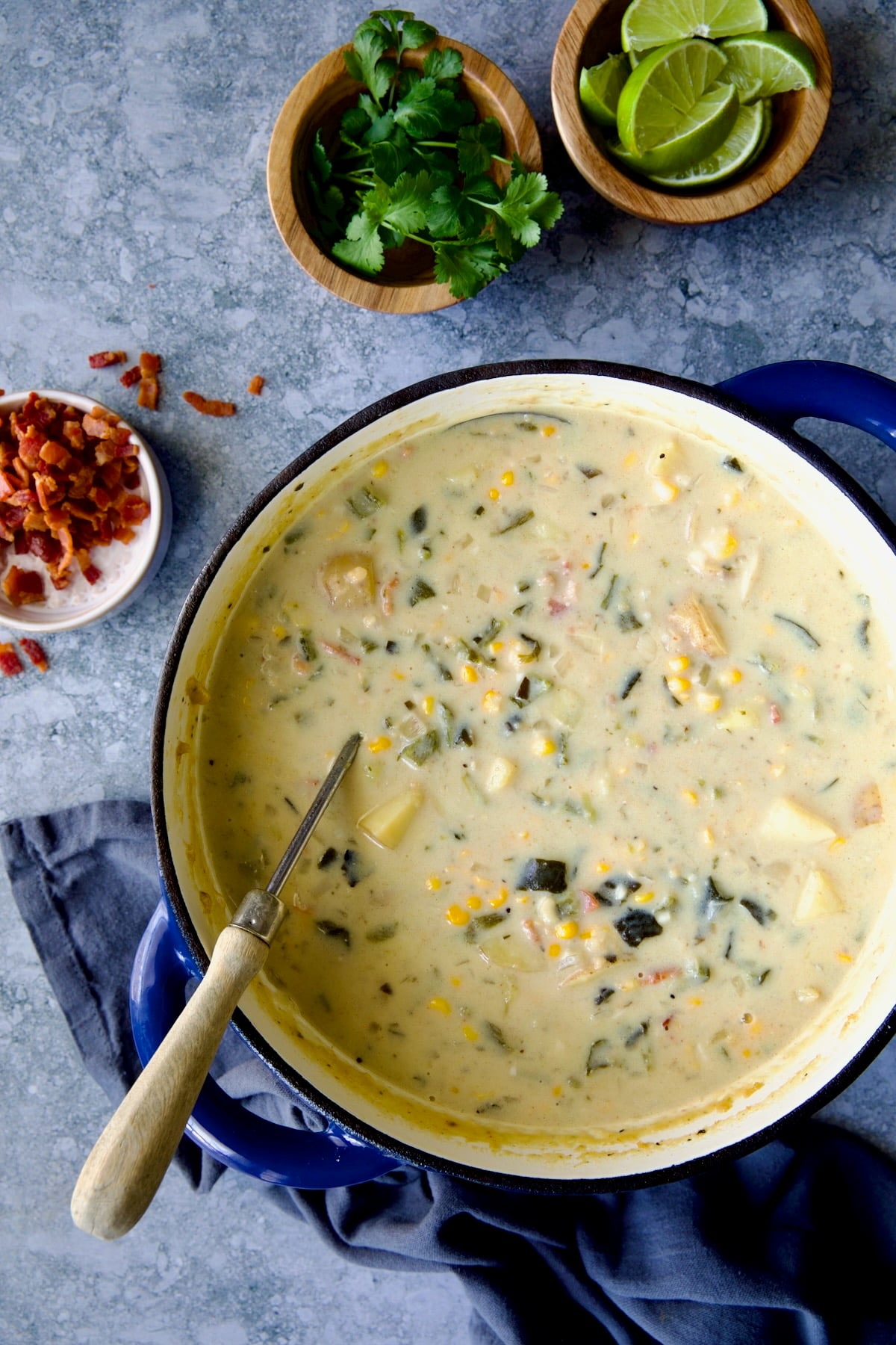 Finished Roasted Poblano Corn Chowder in blue Dutch oven with soup ladle bacon, cilantro and lime wedges in small bowls.