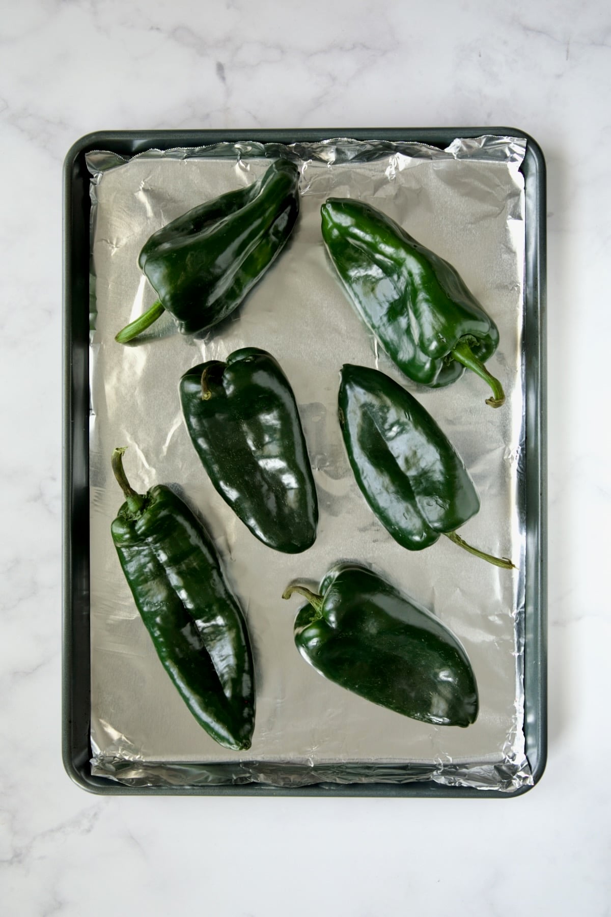 Six fresh, unroasted poblano peppers on aluminum-foil lined rimmed baking sheet.