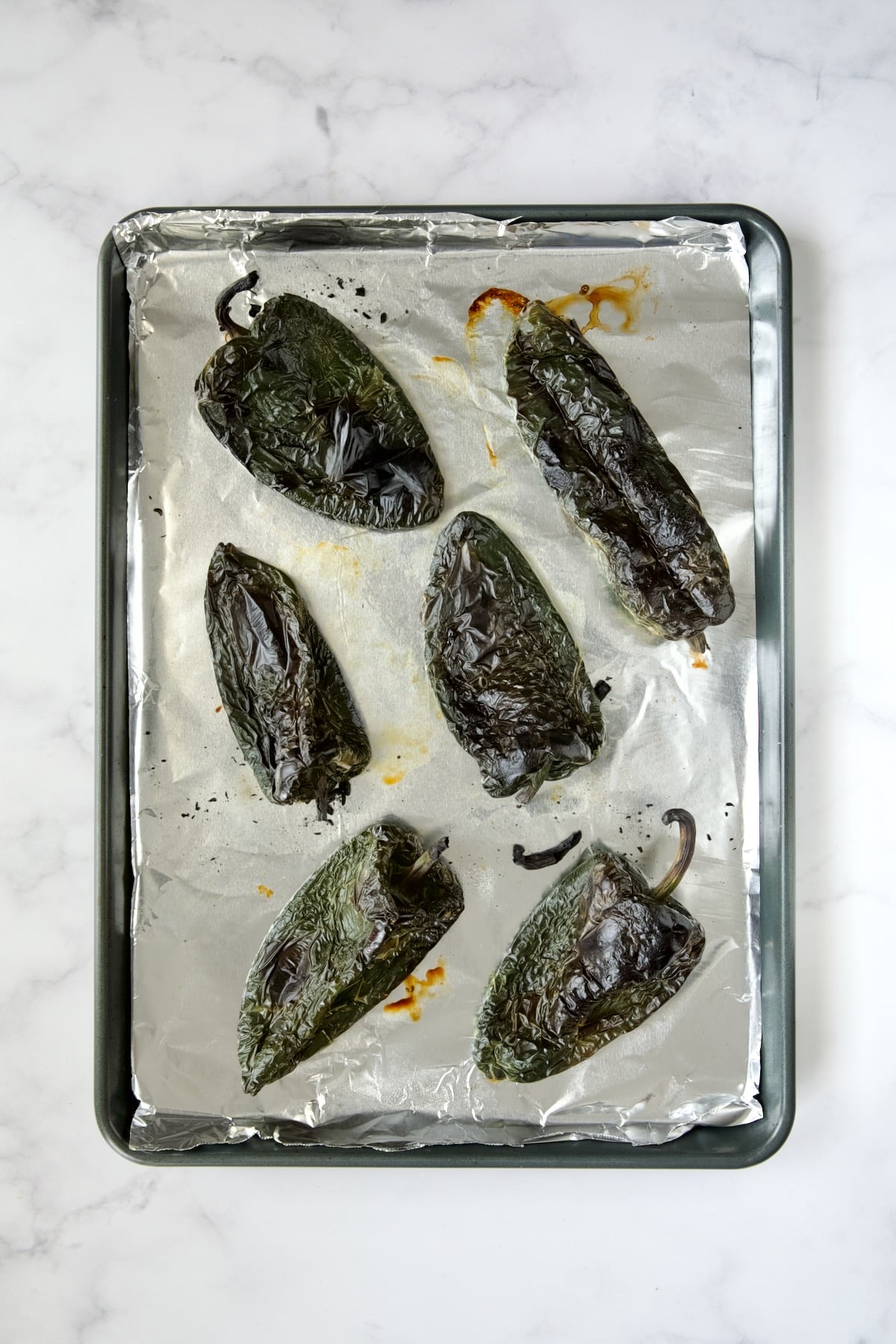 Six roasted poblano peppers on aluminum foil lined rimmed baking sheet.