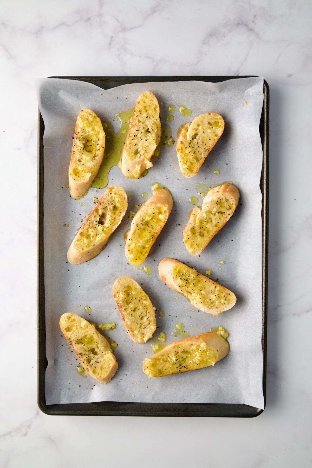 Bread slices on parchment paper lined rimmed baking sheet with olive oil and garlic brushed over the top.