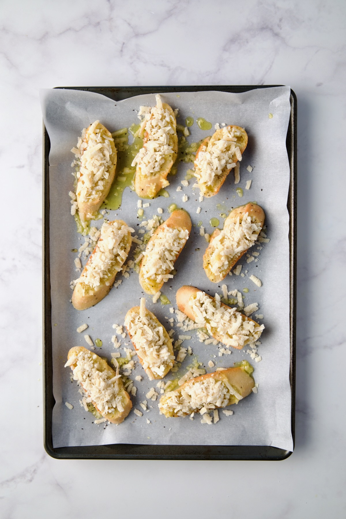 Bread slices with parmesan cheese added on parchment paper lined rimmed baking sheet.