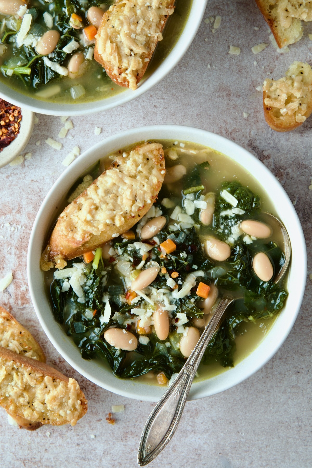 White Bean Kale Soup in white bowl with soup spoon and Parmesan Toast on top.