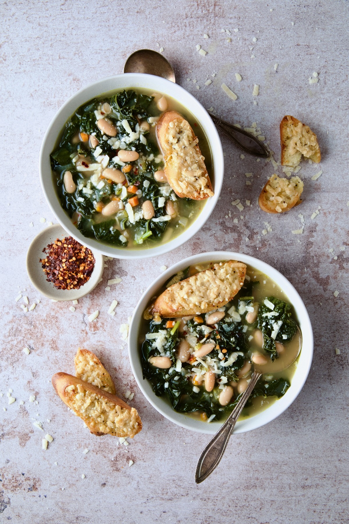 Two bowls of White Bean Kale Soup with Parmesan Toast with crushed red pepper flakes in small bowl.