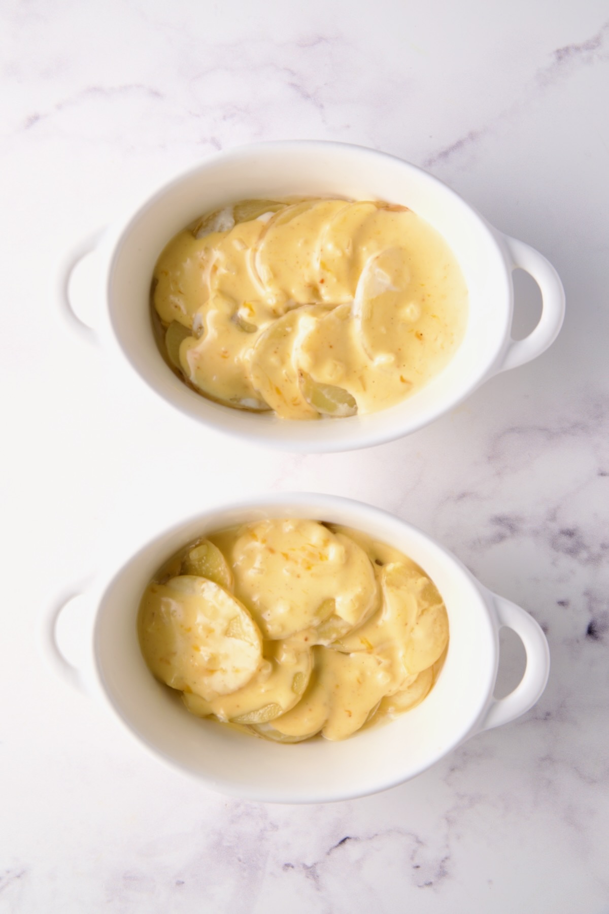 Two oval dishes with first layer of potatoes and cheese.