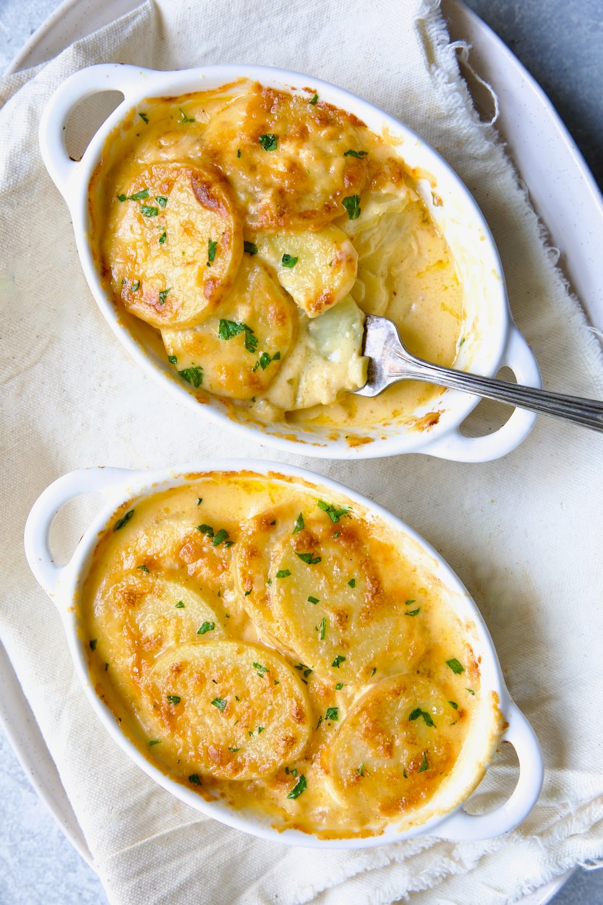 Two white oval dishes of Au Gratin Potatoes for Two with fork digging into one.