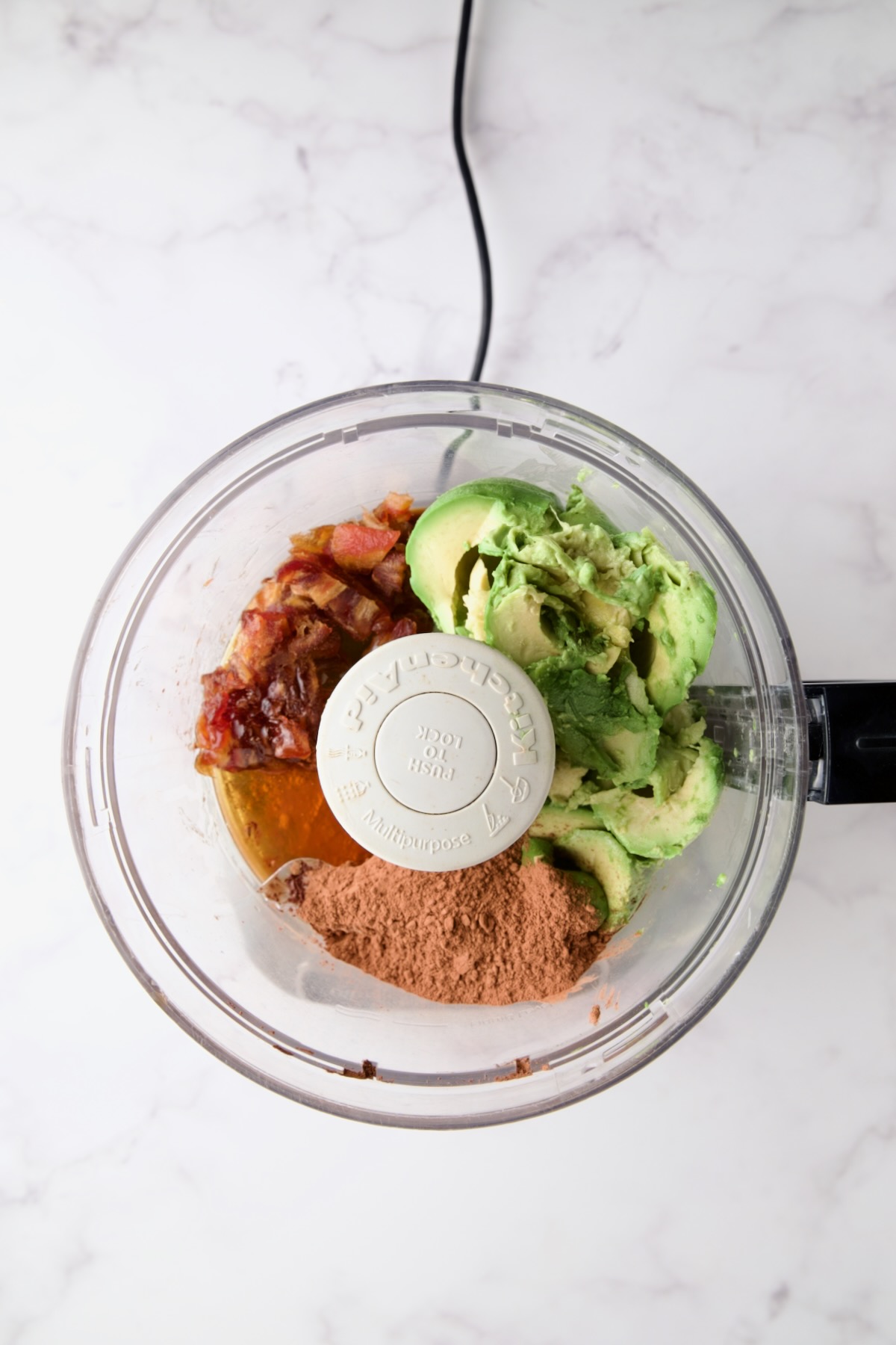 Ingredients for Chocolate Avocado Mousse in food processor before being pureed: Avocado, cocoa powder, Medjool dates, honey and vanilla.
