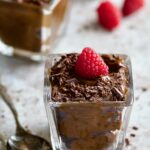 Chocolate Avocado Mousse in square dessert glass with raspberry and chocolate shavings with two spoons.