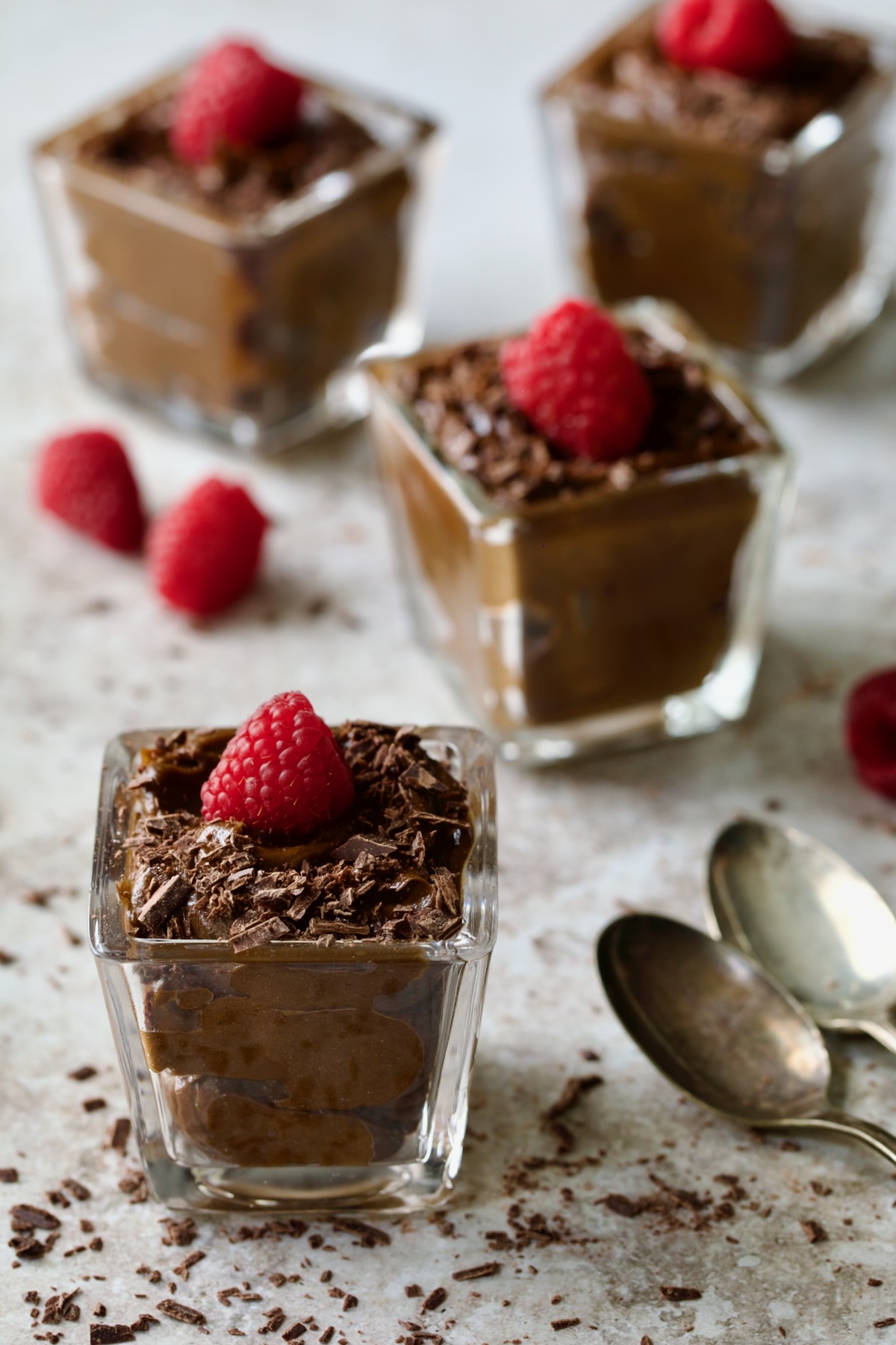 Four square glasses of Chocolate Avocado Mousse garnished with fresh raspberries.