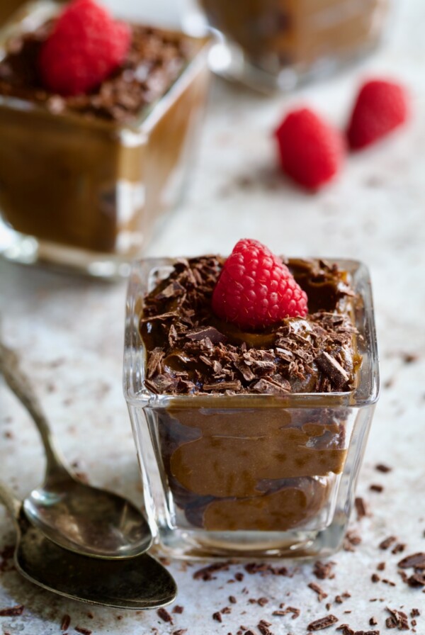 Chocolate Avocado Mousse in square dessert glass with raspberry and chocolate shavings with two spoons.