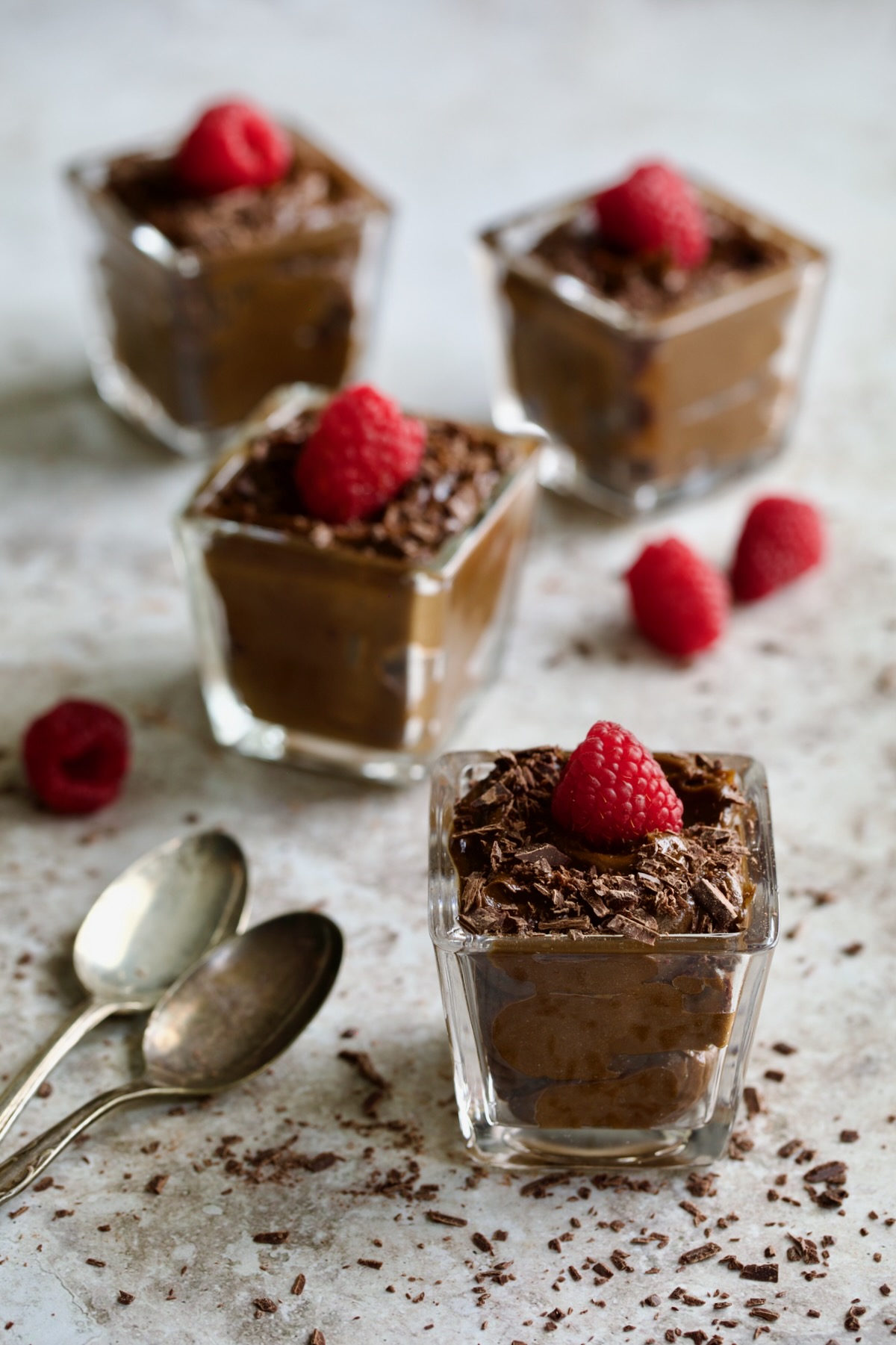Four square glasses of Chocolate Avocado Mousse garnished with fresh raspberries.