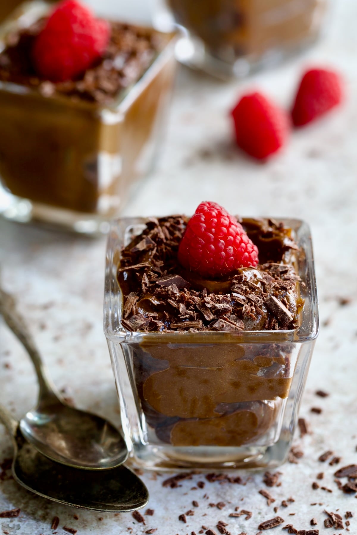 Chocolate Avocado Mousse in square dessert glass with raspberry and chocolate shavings with two spoons.