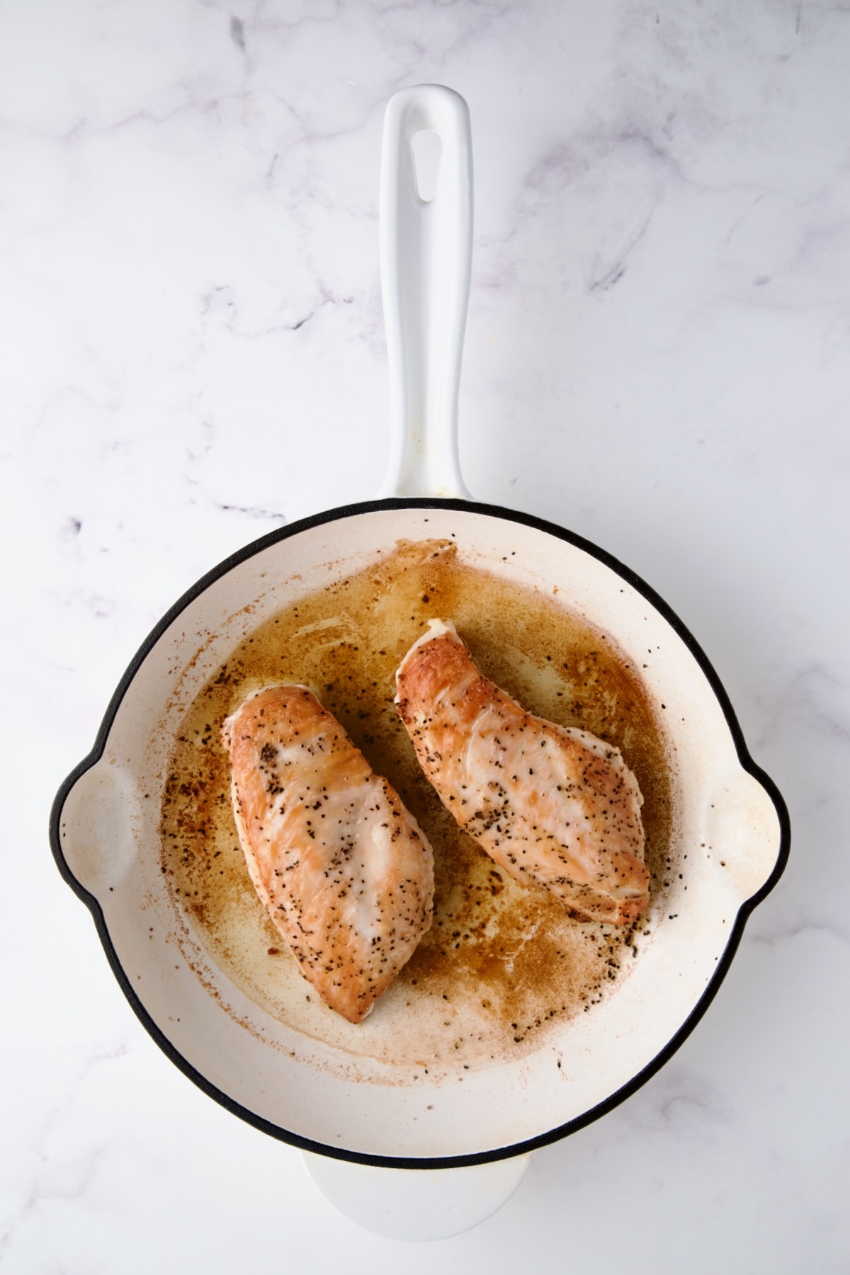 Two browned boneless skinless chicken breast halves in white enamel cast iron skillet.