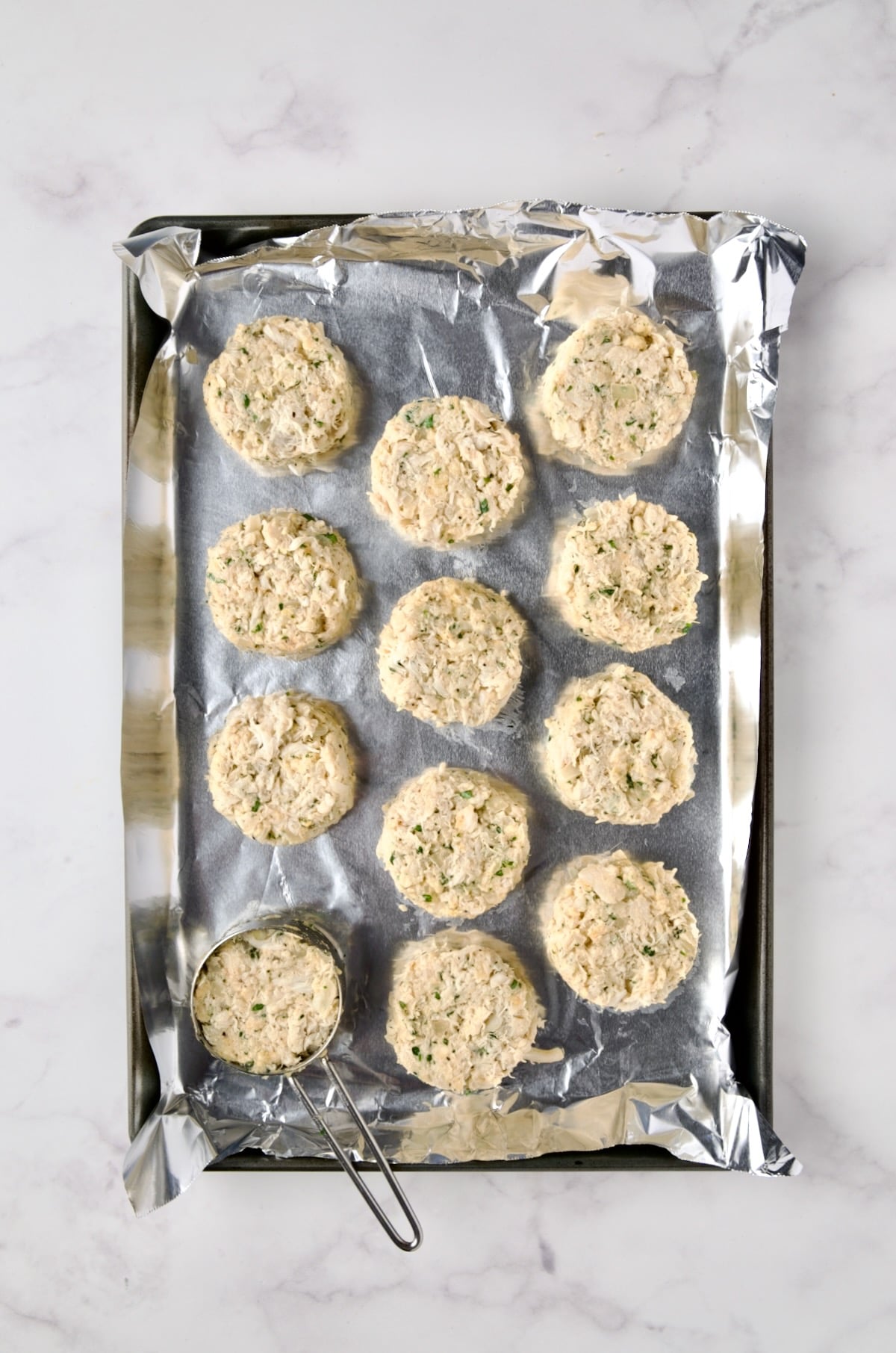 Crab cakes being portioned out on aluminum foil-lined rimmed baking sheet.