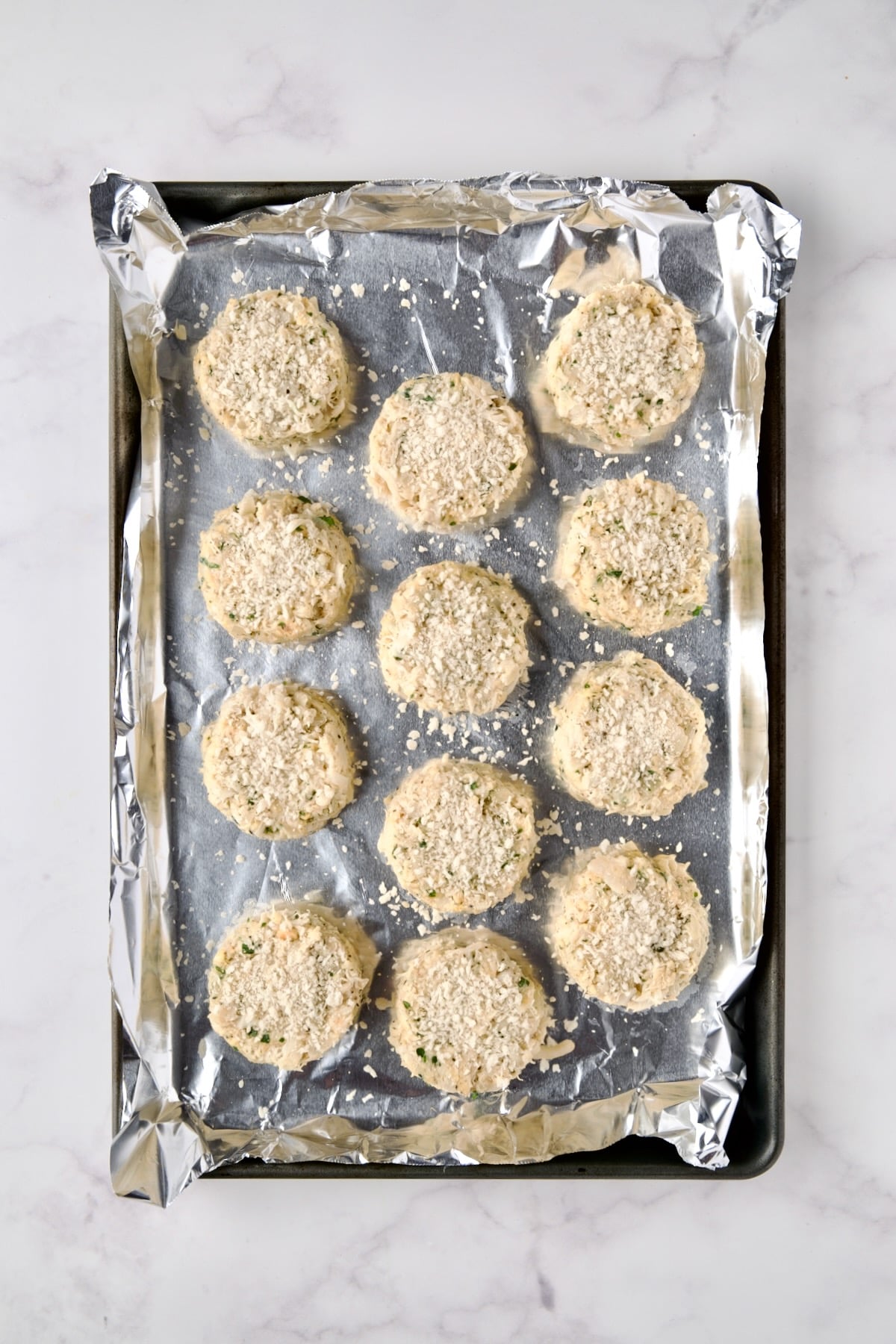 12 crab cakes on aluminum-foil lined rimmed baking sheet with panko sprinkled over the top.