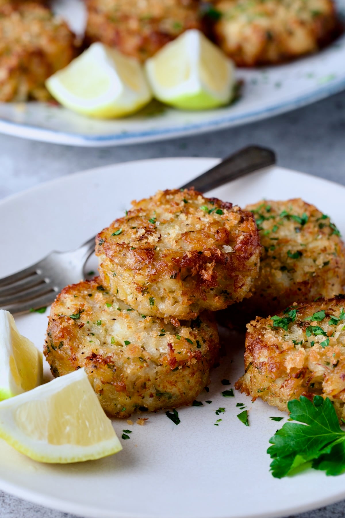 Four Maryland-Style Crab Cakes on white plate with lemon wedges and fork in the background.