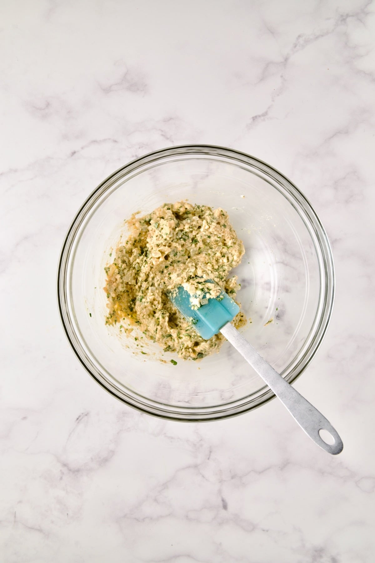 Blue spatula stirring ingredients in glass bowl for crab cakes before crab is added.