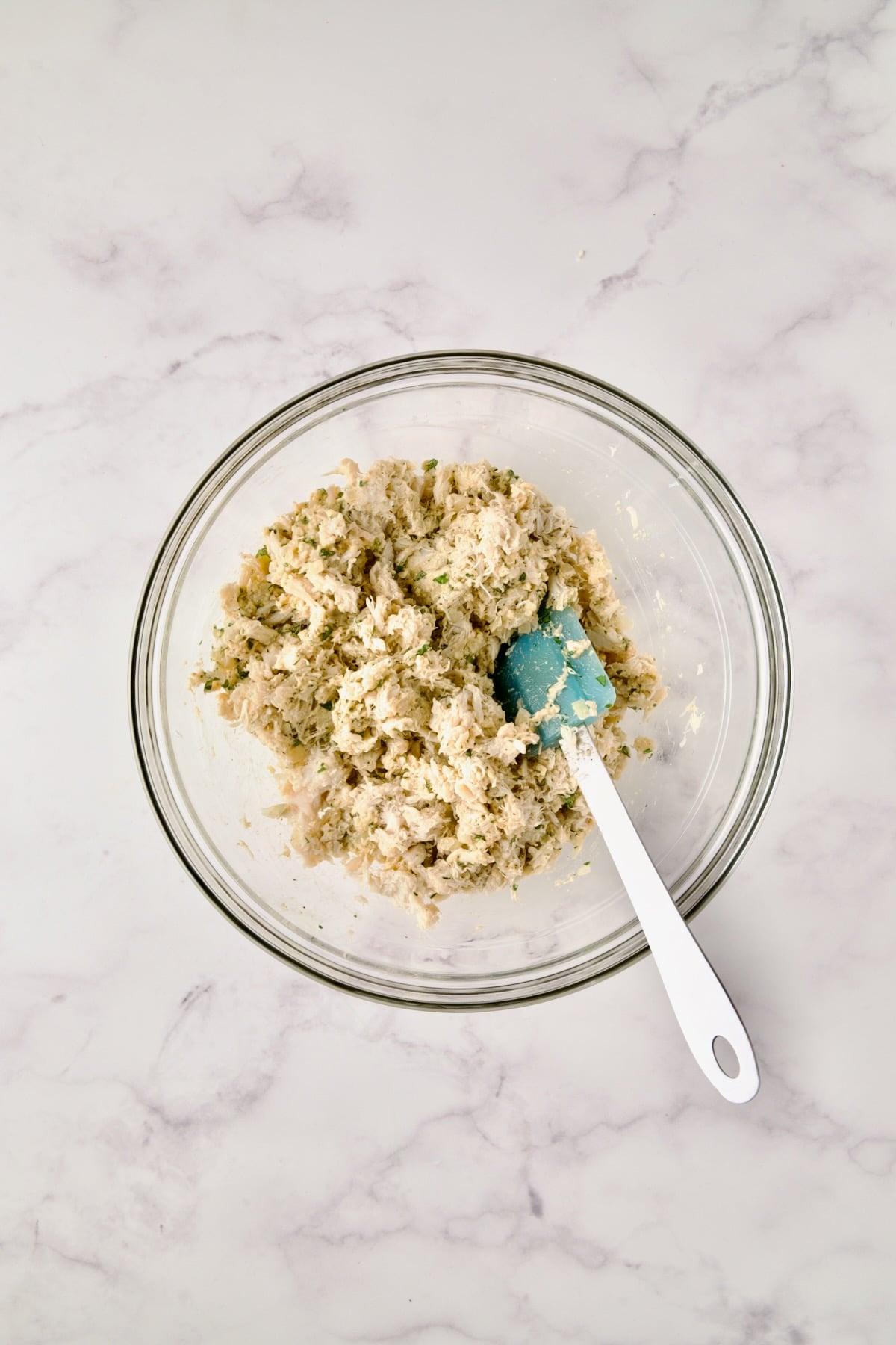 Blue spatula stirring all the ingredients for crab cakes in glass bowl.