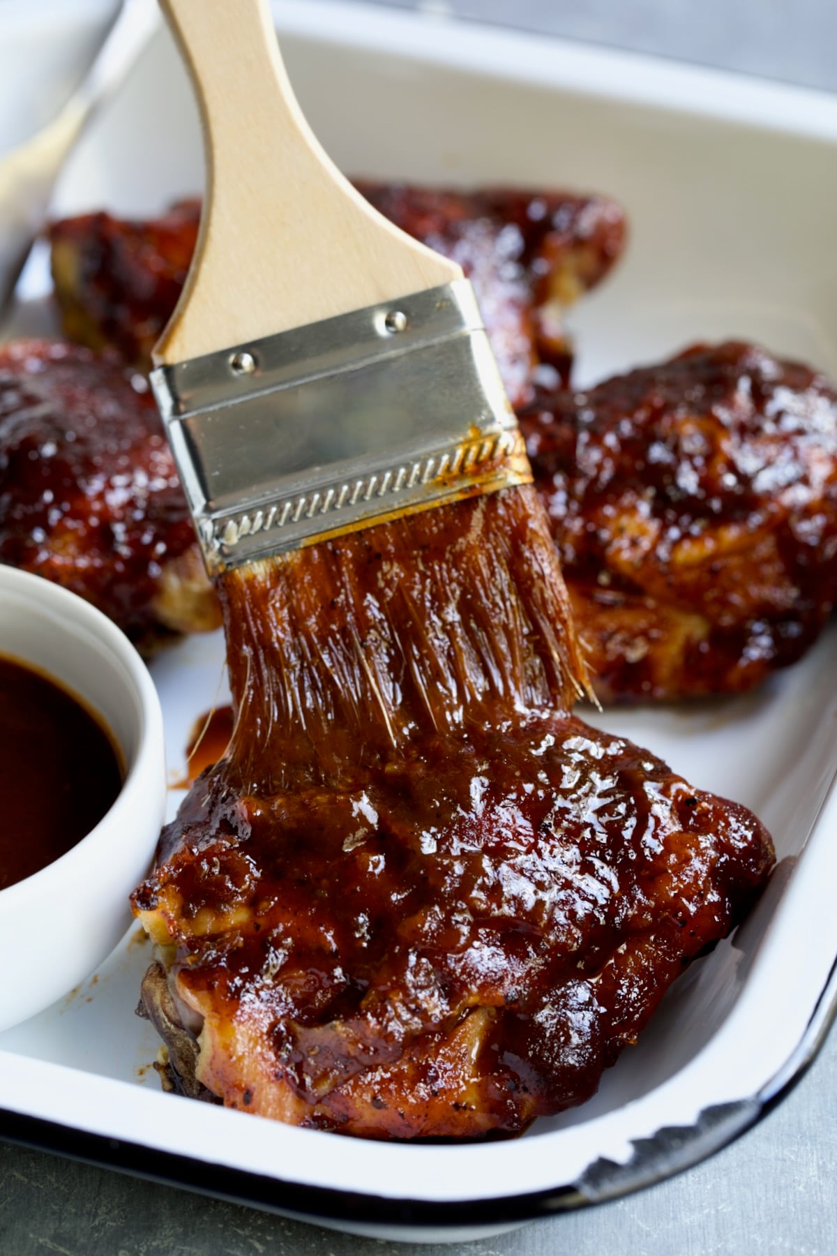 Baked BBQ Chicken Thigh being brushed with sauce in white enamel serving dish.