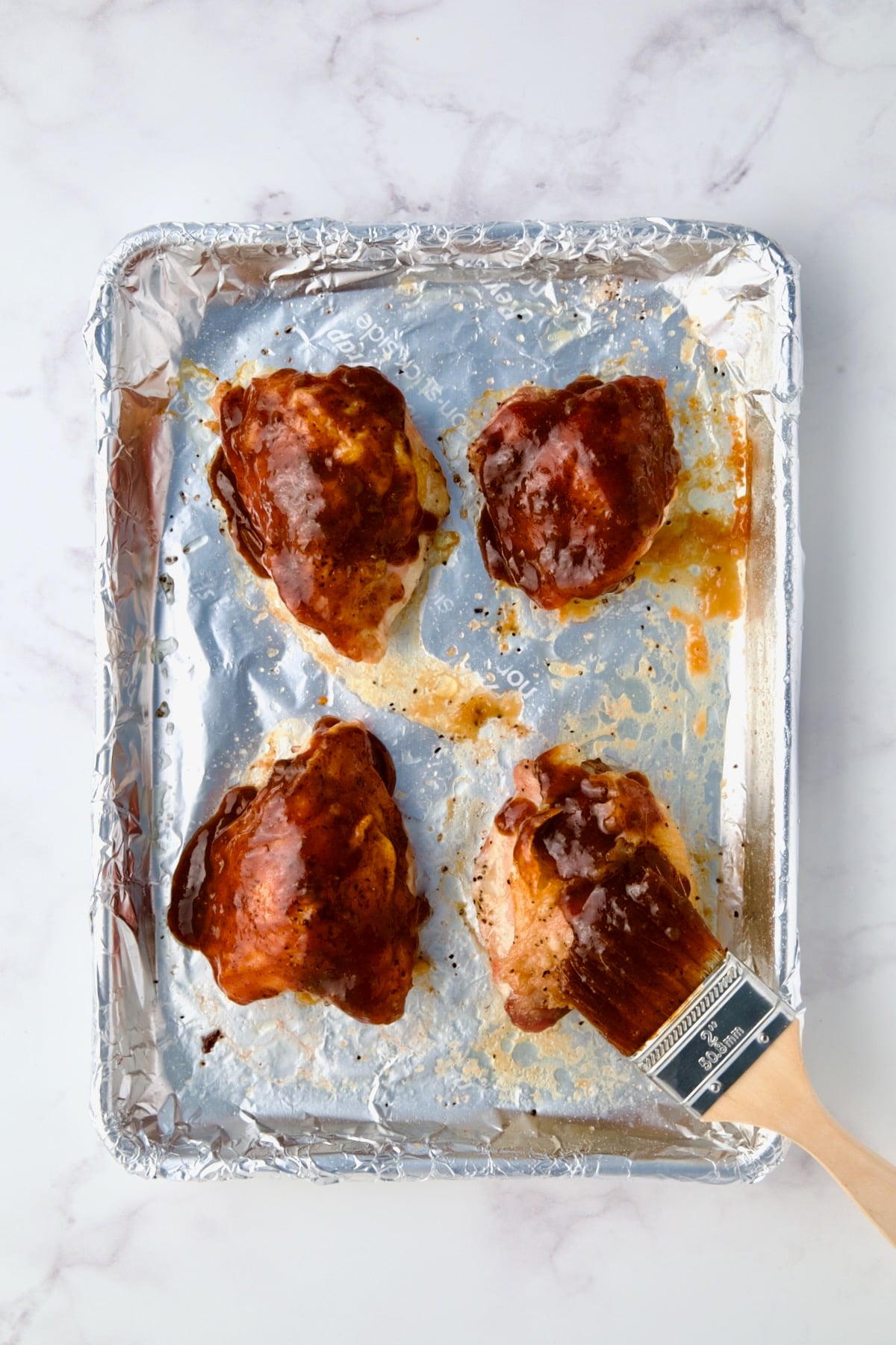 Four chicken thighs being brushed with barbecue sauce on aluminum foil-lined baking sheet.