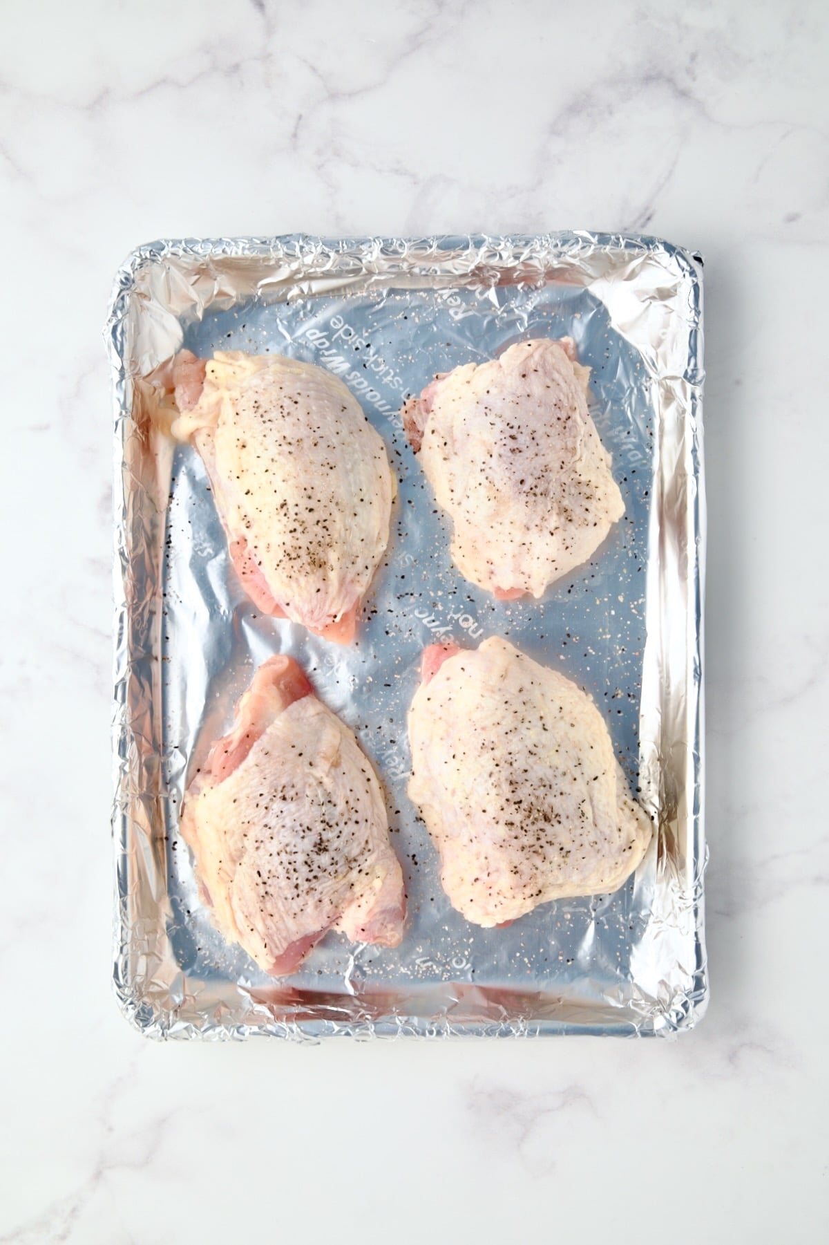 Four uncooked chicken thighs on aluminum foil-lined baking sheet.