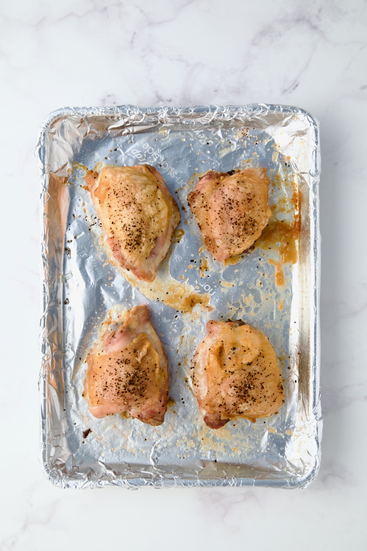 Four baked chicken thighs on aluminum foil-lined baking sheet.