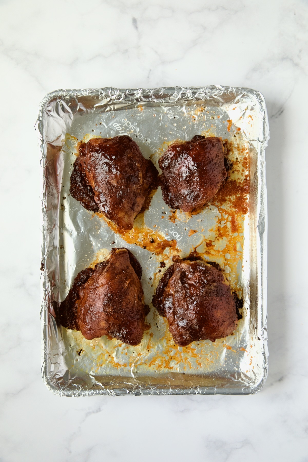 Four finished barbecue chicken thighs on aluminum foil-lined pan.