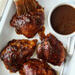 Four Baked BBQ Chicken Thighs in white enamel serving tray with bowl of barbecue sauce.