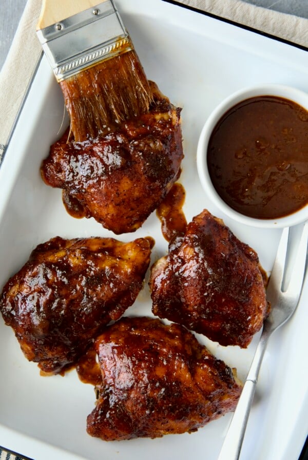 Four Baked BBQ Chicken Thighs in white enamel serving tray with bowl of barbecue sauce.