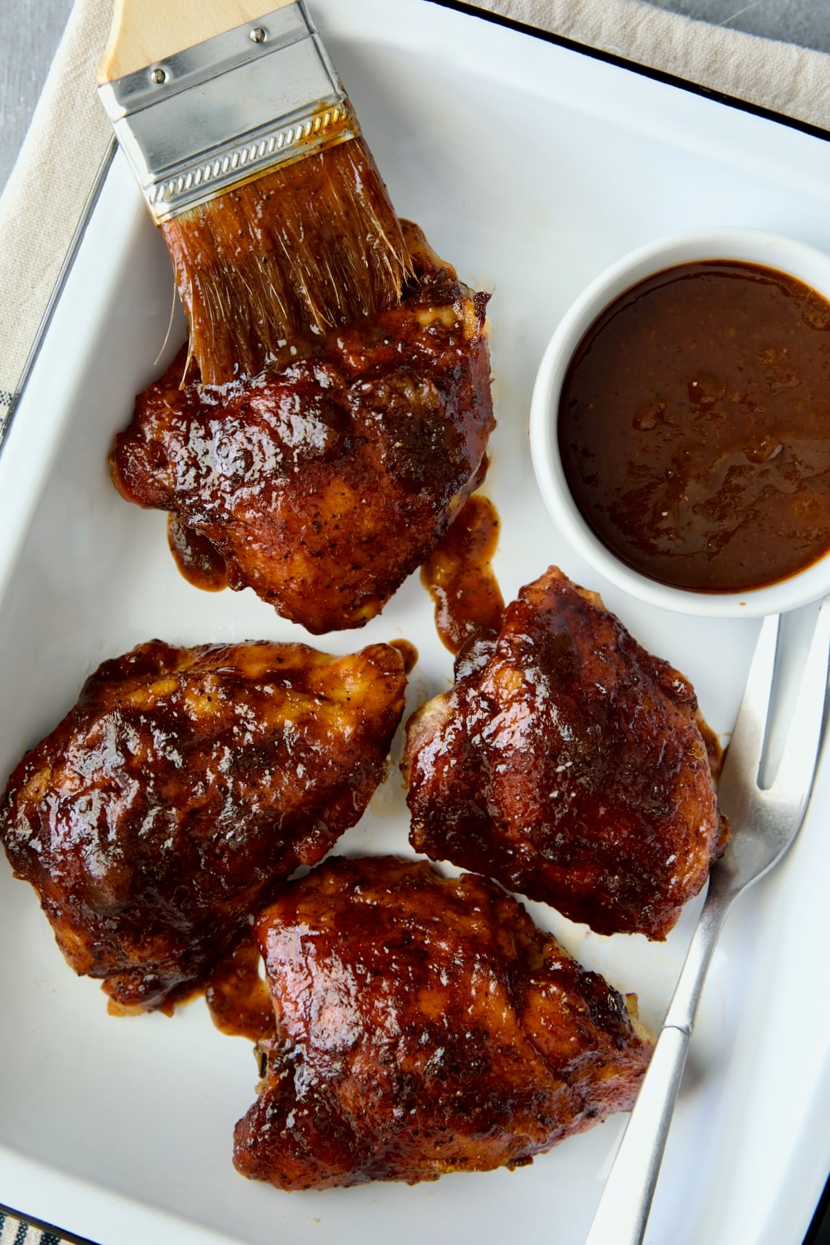 Four Baked BBQ Chicken Thighs in white enamel serving tray with bowl of barbecue sauce.