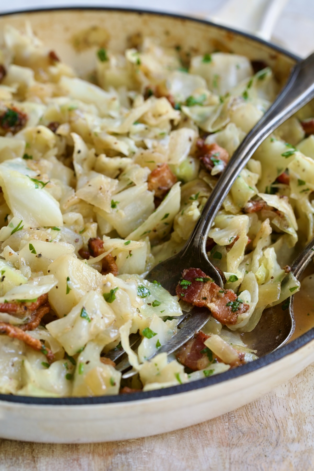 Finished Fried Cabbage with Bacon in white enamel cast iron skillet with serving utensils.