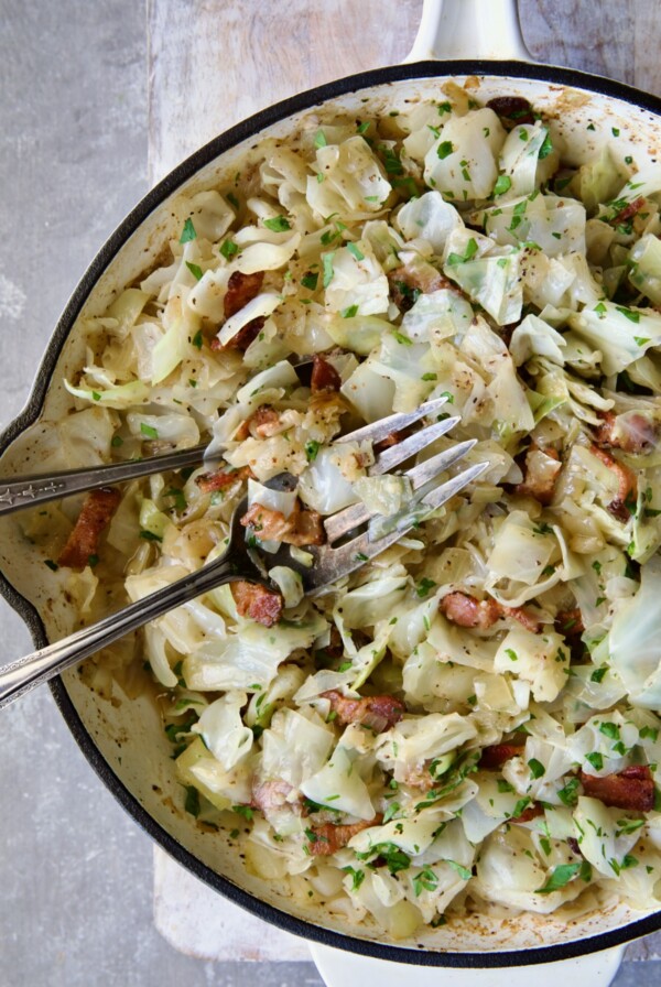 Two serving utensils serving up Fried Cabbage with Bacon from white enamel cast iron skillet.