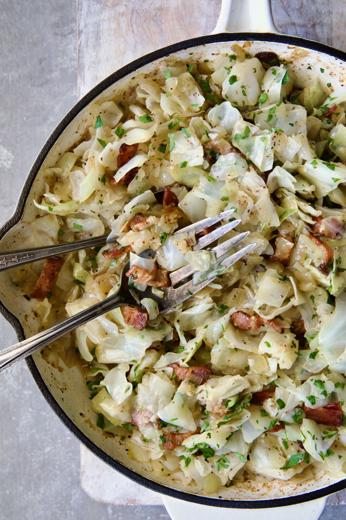 Two serving utensils serving up Fried Cabbage with Bacon from white enamel cast iron skillet.
