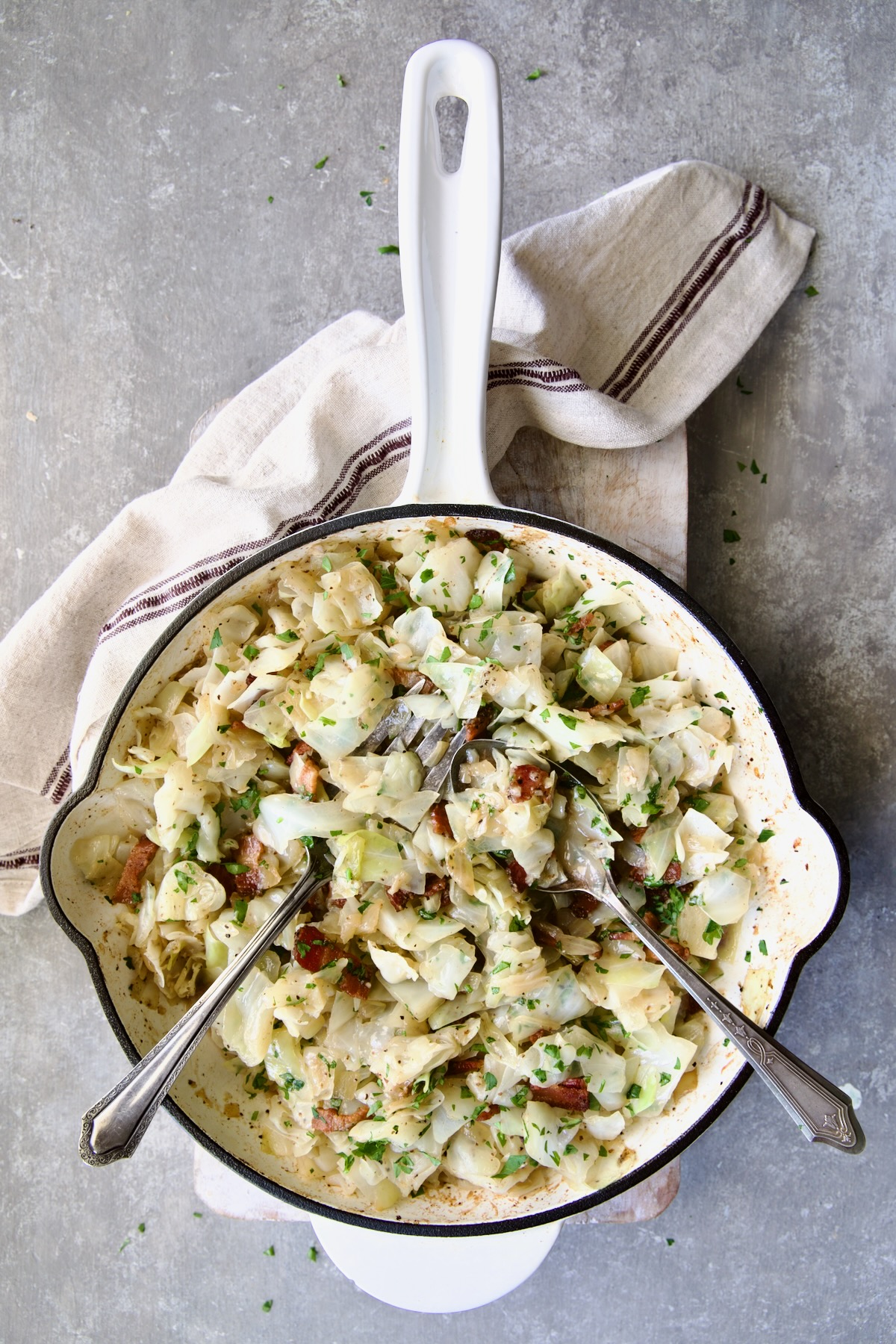 Fried Cabbage with Bacon in white enamel cast iron skillet with serving utensils on white cutting board with red striped napkin.