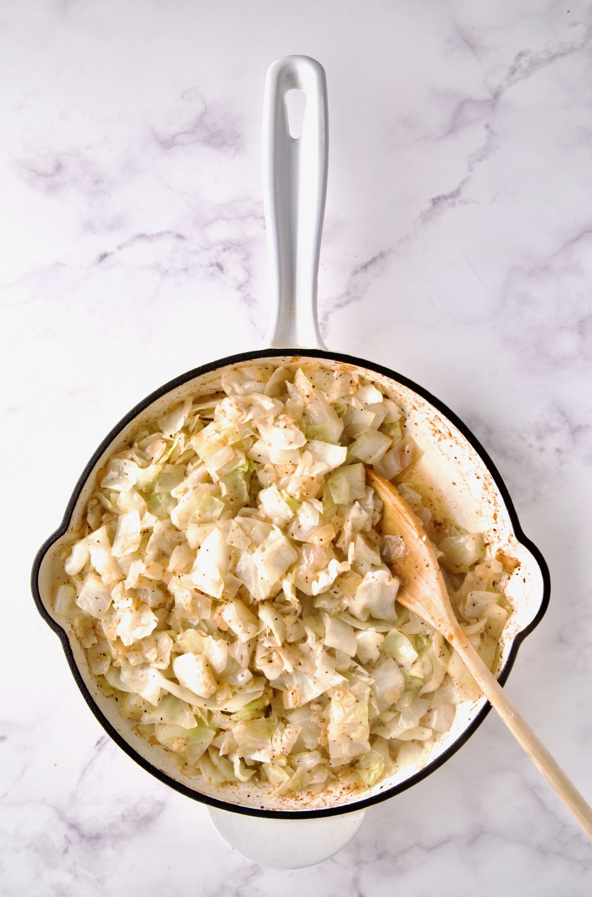 Wooden spoon stirring cabbage after chicken broth is added in white enamel cast iron skillet.