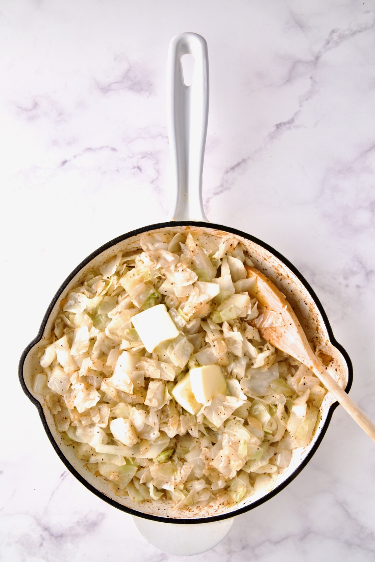 Butter being stirred into cabbage in white enamel cast iron skillet with wooden spoon.