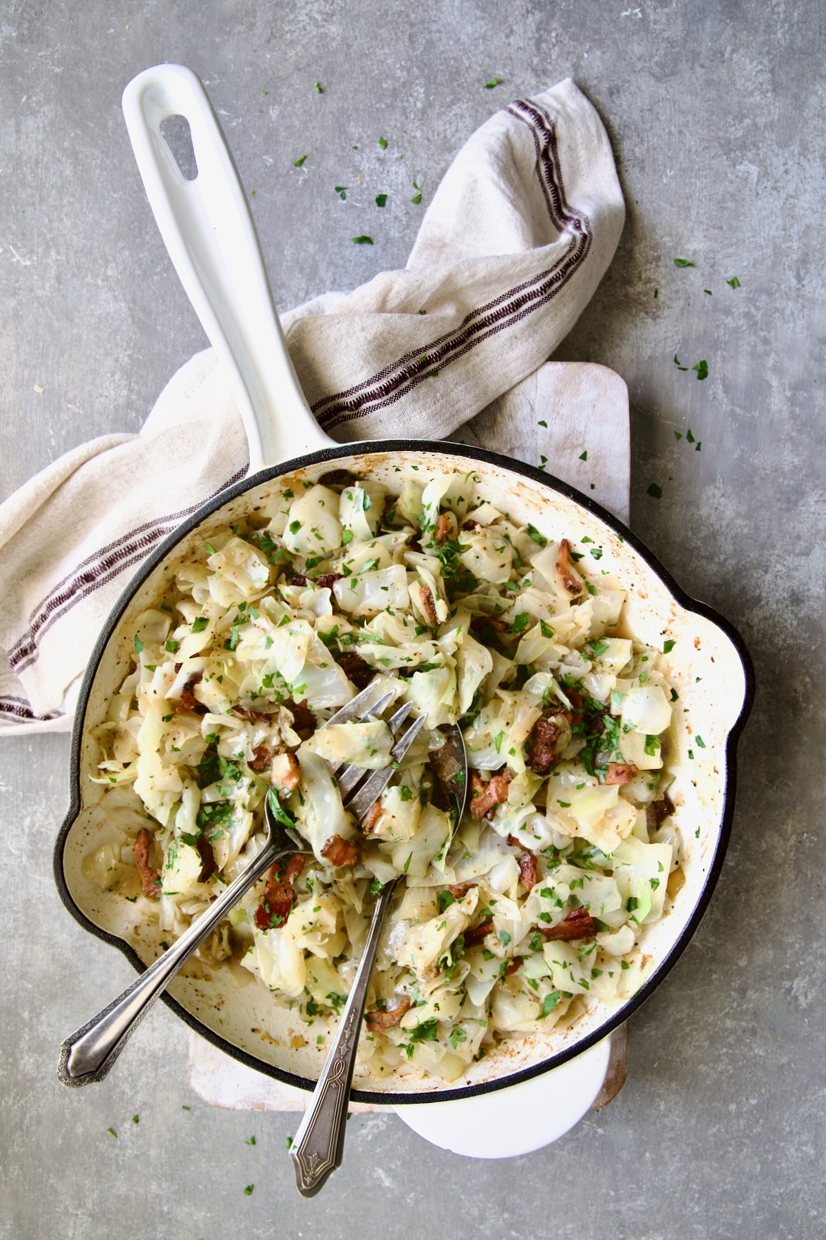 Fried Cabbage with Bacon in white enamel cast iron skillet with serving utensils and red striped napkin.