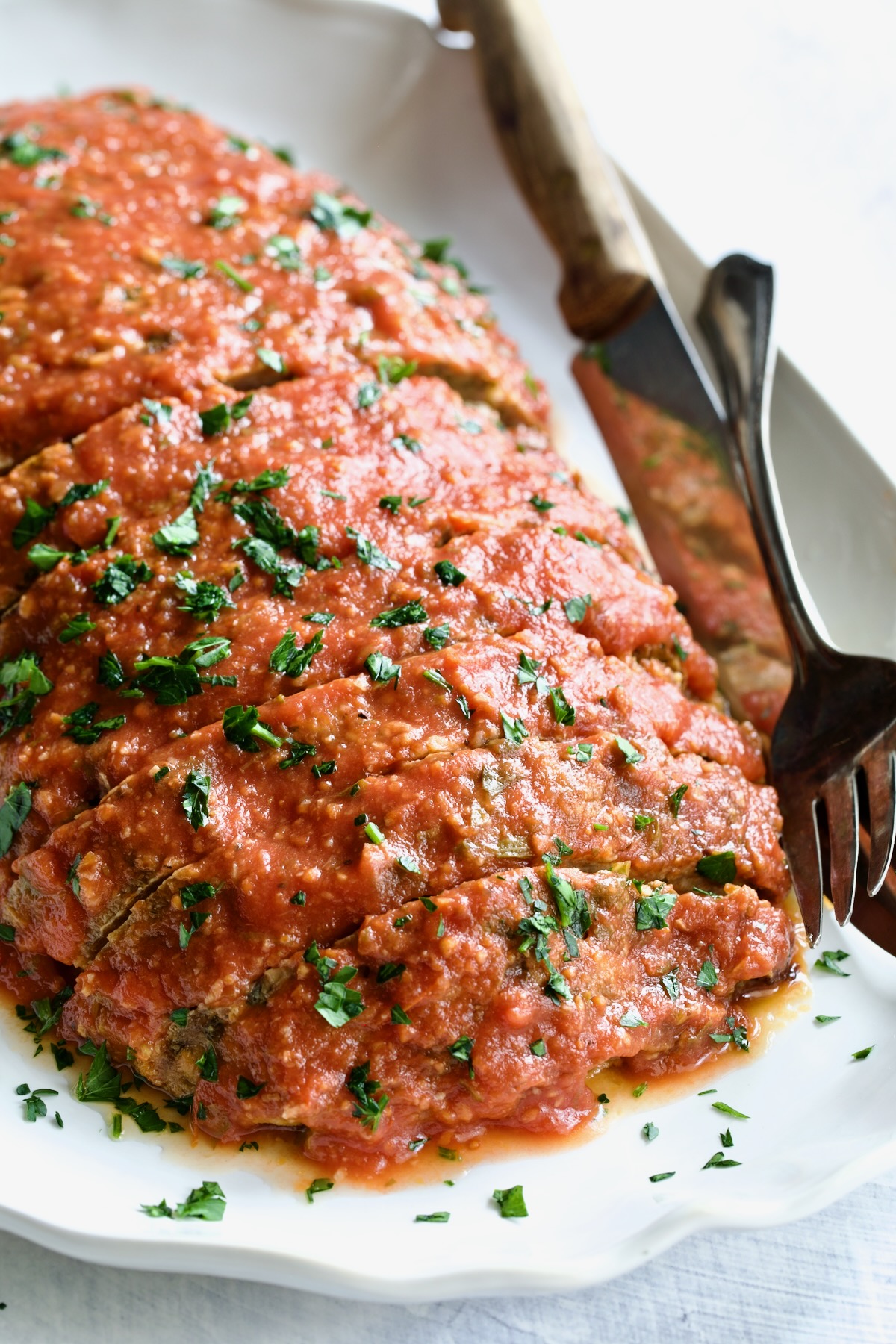 Italian Meatloaf on white platter garnished with fresh parsley with serving utensils.